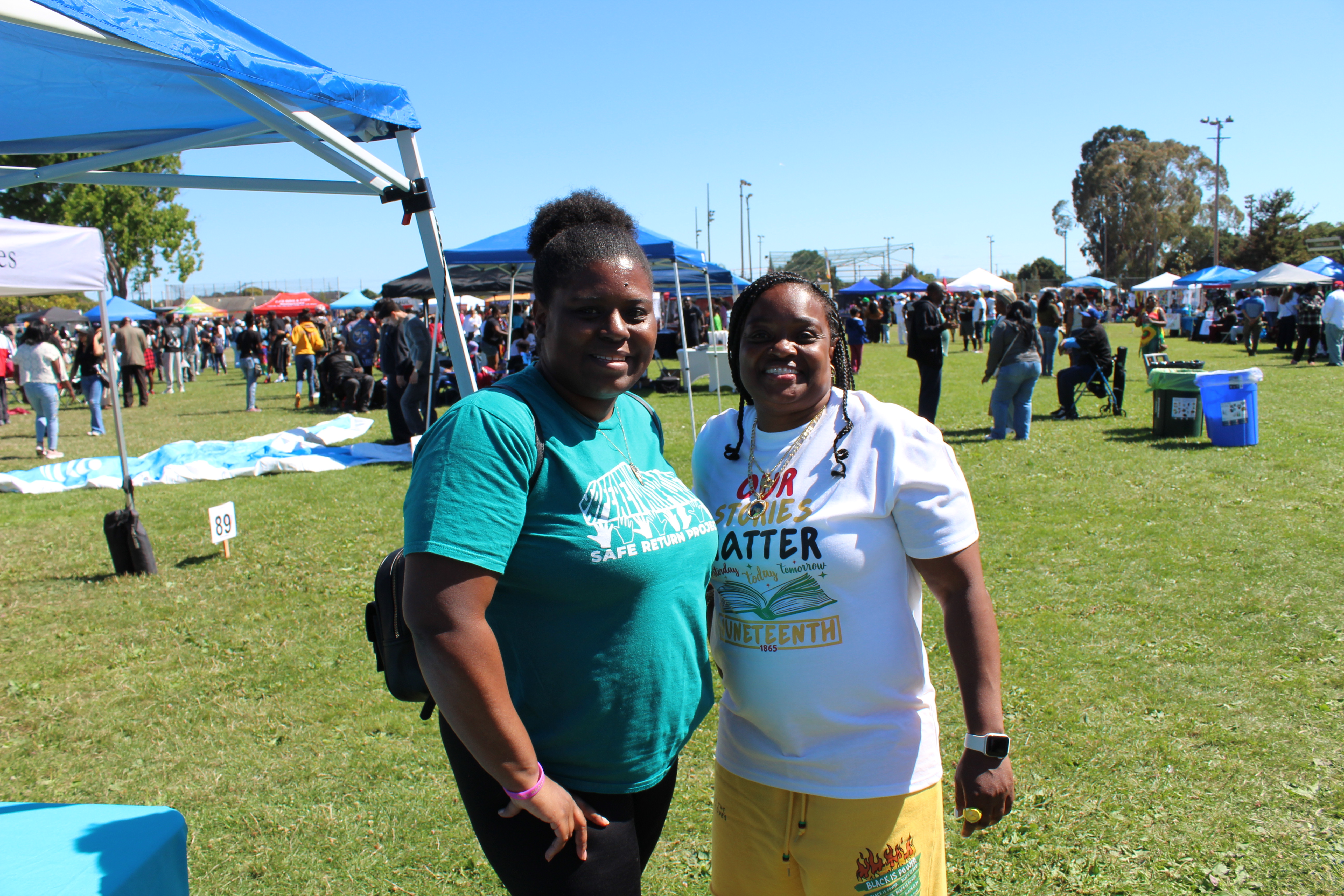 Two women smiling together at a Juneteenth community celebration in a park, wearing Safe Return Project and 'Our Stories Matter' shirts.